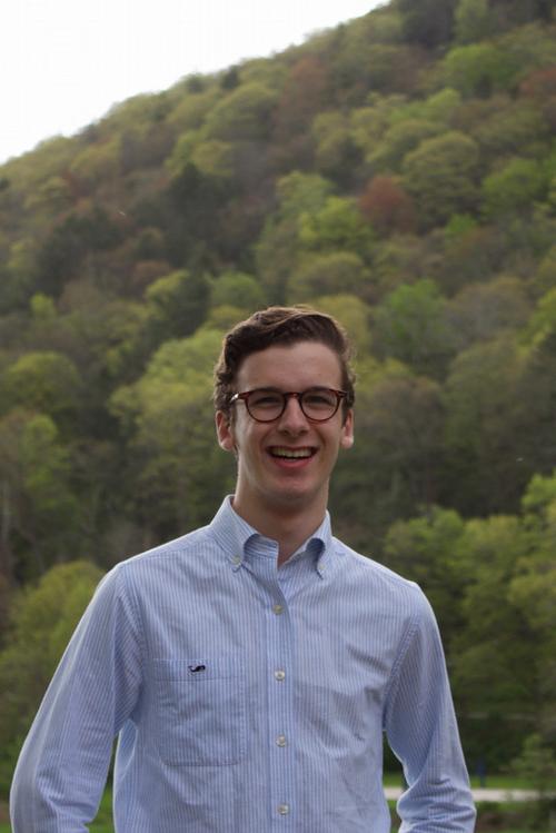 布兰登·舒斯特微笑的照片, 穿着一件浅蓝色的钮扣衬衫, 背景是绿树成荫的山坡